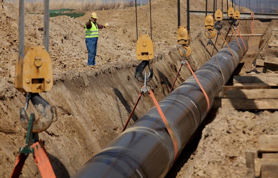 Se observa una tubería de Diámetros Mayores siendo instalada en una excavación, con un operario utilizando equipo de seguridad que da instrucciones mientras la tubería es levantada con grúas. El entorno es de construcción, con tierra y maquinaria alrededor.