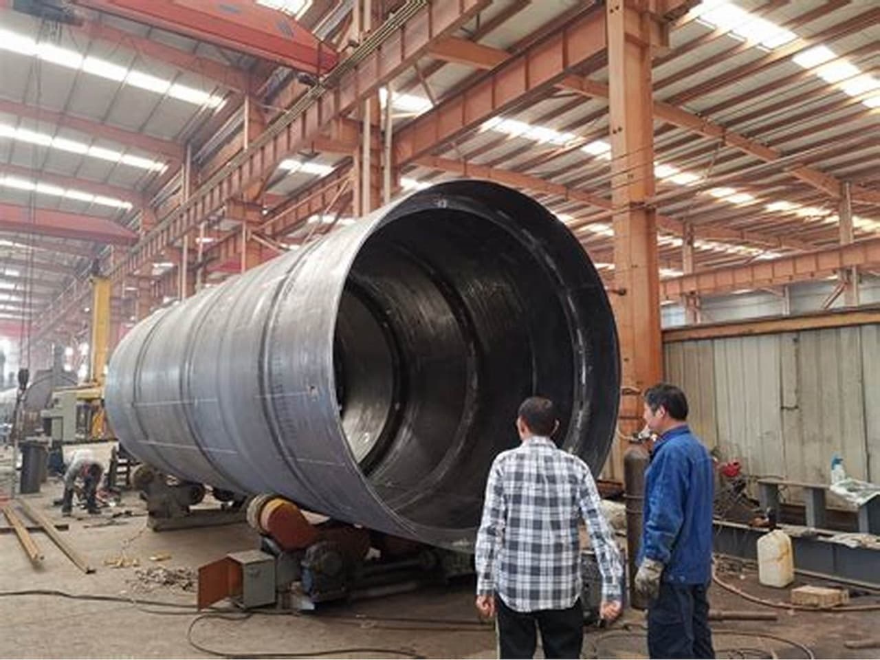 En un taller industrial, se observa una tubería de Diámetros Mayores de gran tamaño, con trabajadores que la inspeccionan. El ambiente es amplio, con maquinaria y herramientas a la vista, y una estructura metálica en el techo.