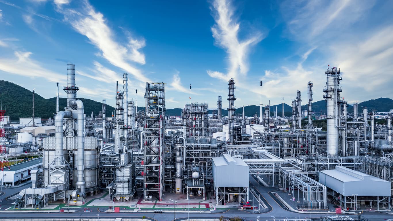Instalaciones industriales complejas con estructuras de Tubería de Diámetros Mayores, torres de destilación y maquinaria, todo bajo un cielo azul con nubes.