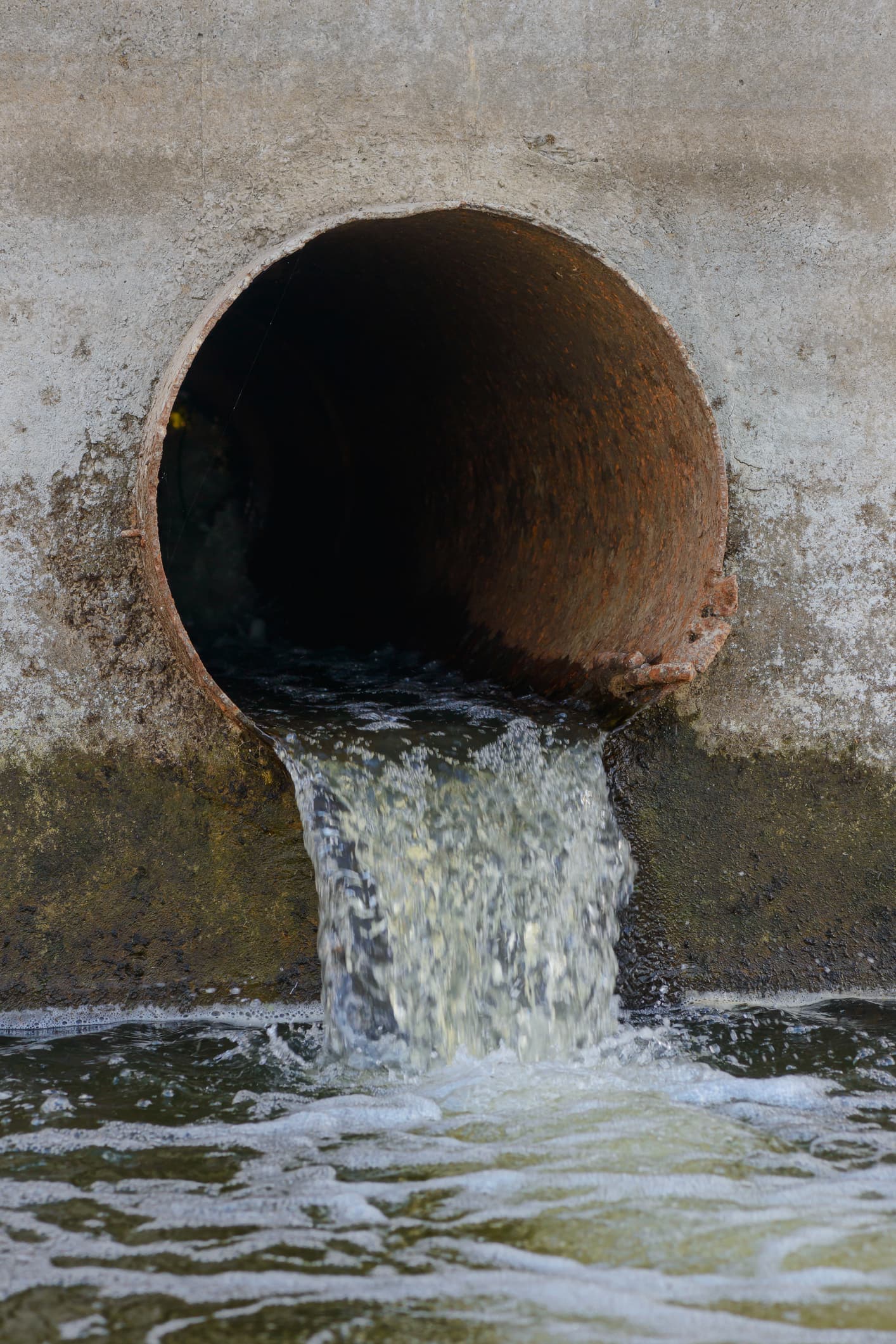 Tubería de Diámetros Mayores de acero que presenta signos de rustificación, desde donde fluye agua con espuma hacia el exterior, mostrando un entorno de concreto y suciedad.